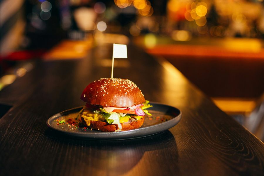 A burger with lettuce, tomato, and cheese on a black plate sits on a dark wooden table in a restaurant.