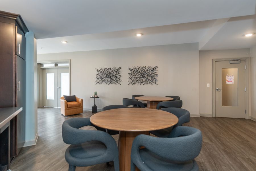 Modern lounge area with round tables, gray chairs, wall art, and an orange armchair near glass doors.