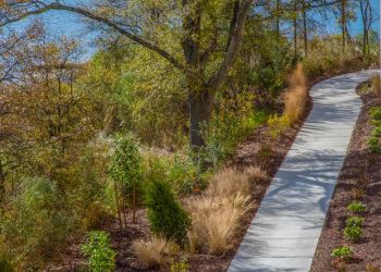 Attain on 5th apartment homes with Curved concrete path with plants and trees beside a blue river on a sunny day.