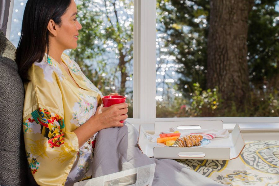 Woman in a robe sits in bed with a red mug, breakfast tray, and newspaper, looking out at a sunny lake view.