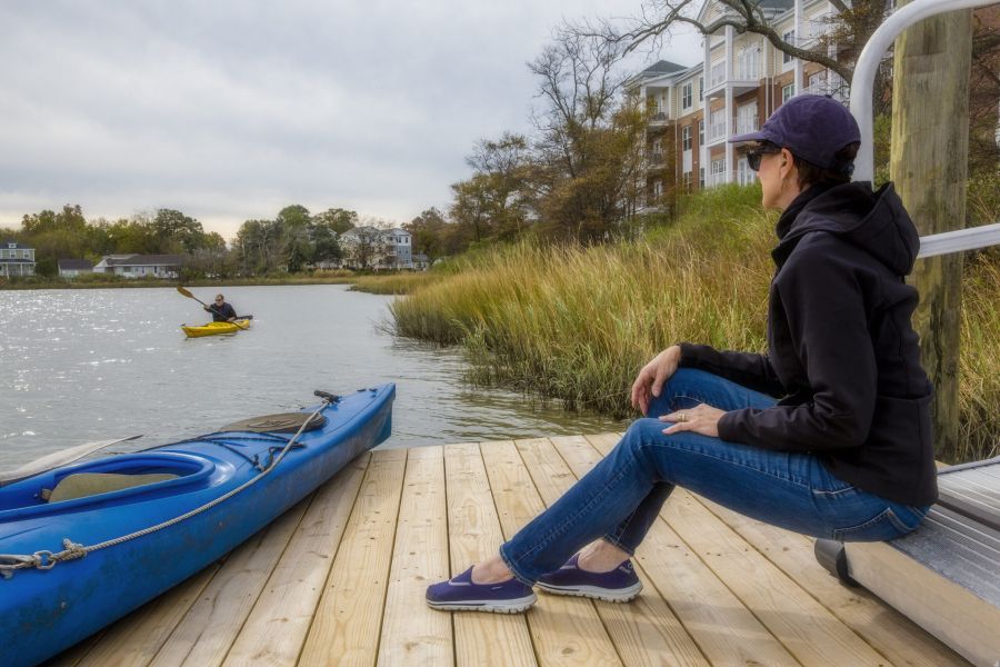 Woman sitting on a dock near a kayak, watching another person paddling on the water by the shore.