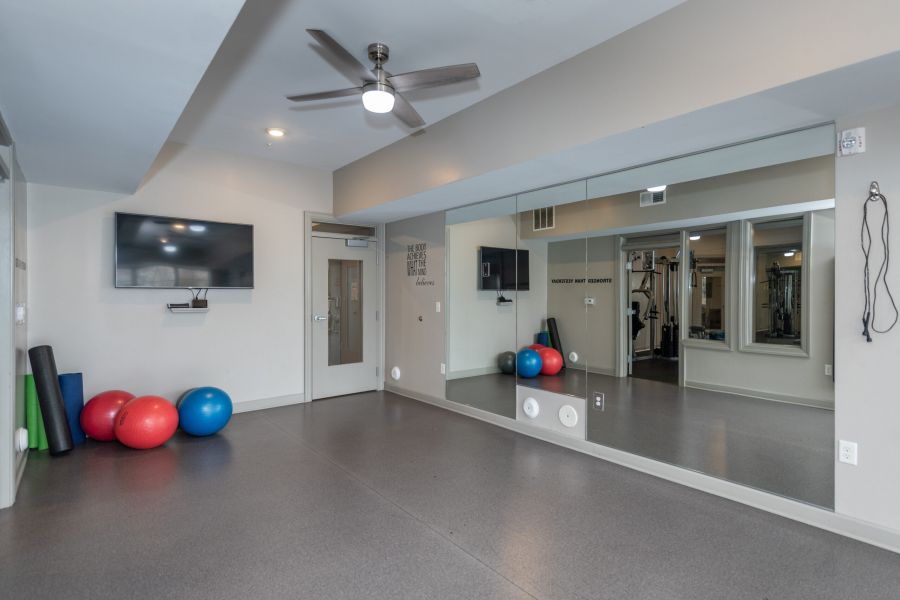 Exercise room with mirrored wall, TV, ceiling fan, yoga mats, and three colorful exercise balls.