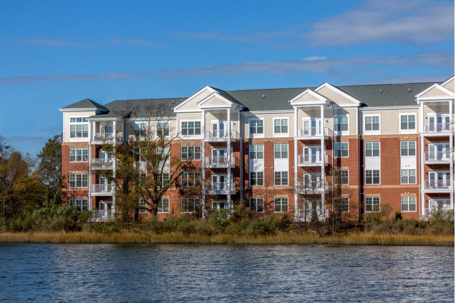 Red brick apartment building with balconies, next to a body of water under a clear blue sky.