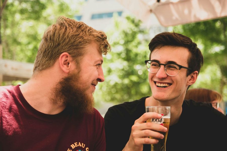 Two young men sit outdoors, smiling and chatting while one holds a glass of beer.