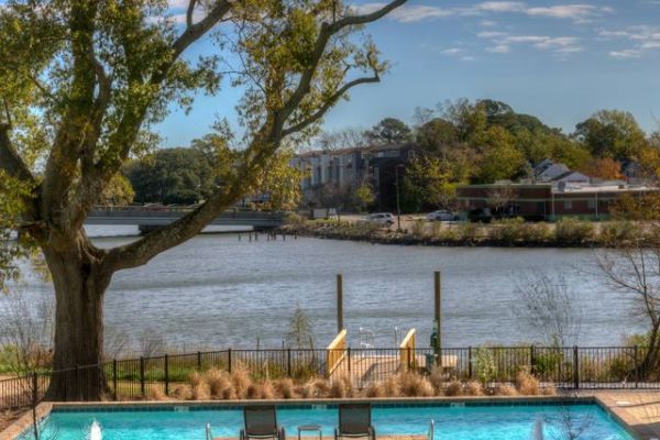 A riverside pool with lounge chairs, trees, and houses visible across the water under a blue sky.