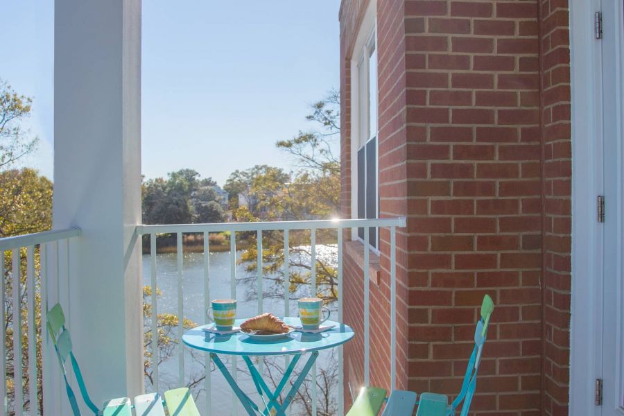 Two turquoise chairs and a table with drinks and food on a sunny balcony overlooking water and trees.