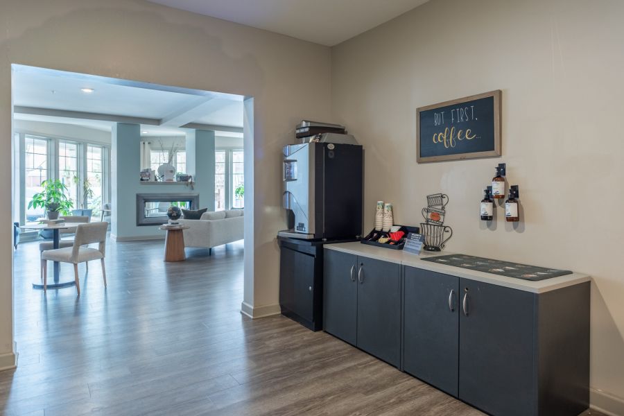 Coffee station with supplies on cabinets, a small fridge, and a sign reading "But first, coffee" by a lounge area.