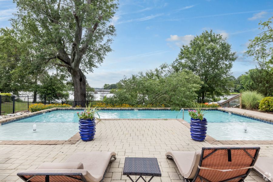 Two lounge chairs face a pool with blue planters, surrounded by trees and overlooking a lake.